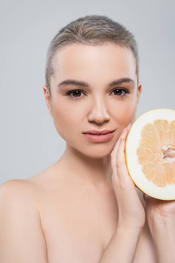портрет молодой женщины с половиной сочного грейпфрута, смотрящей на камеру, изолированную на сером - Фото, изображение