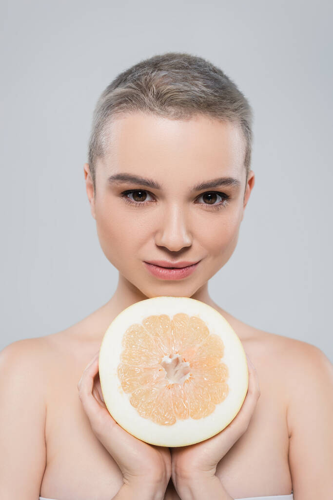 junge Frau mit nackten Schultern blickt in die Kamera, fast die Hälfte der Grapefruit isoliert auf grau - Foto, Bild