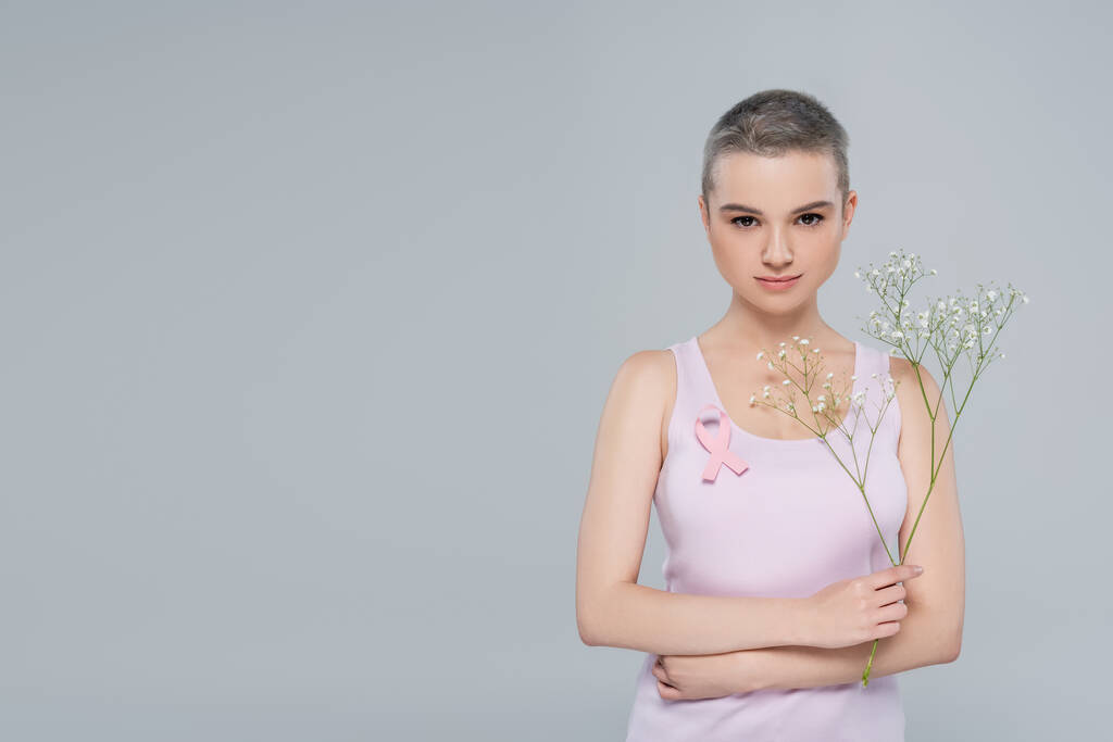νεαρή γυναίκα με κορδέλα ευαισθητοποίησης για τον καρκίνο του μαστού και γυψόφυλλες λουλούδια που απομονώνονται σε γκρι - Φωτογραφία, εικόνα