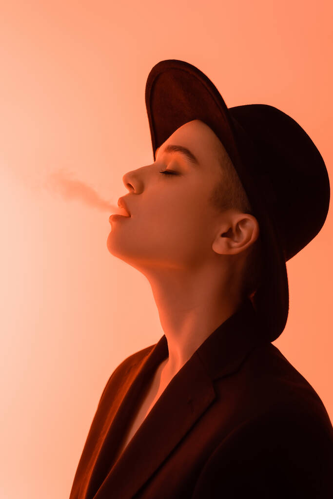 vista laterale di giovane donna in cappello tesa nera espirando fumo su sfondo corallo - Foto, immagini