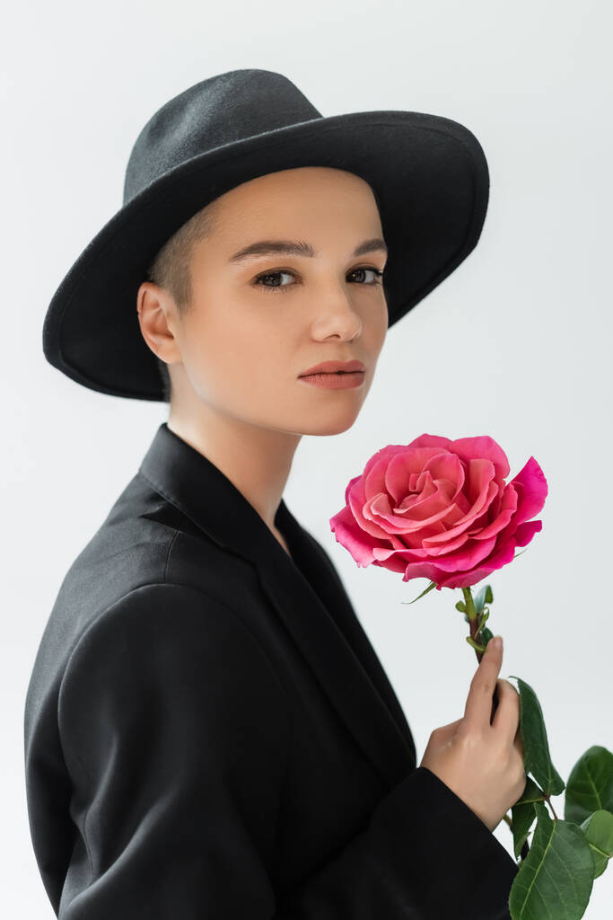 Charmante Frau mit natürlichem Make-up, trägt schwarzen Fedora-Hut, hält rosa Rose isoliert auf grau - Foto, Bild