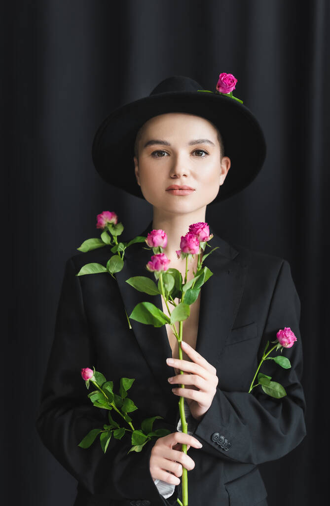 jovem com maquiagem natural, vestindo chapéu de bainha e blazer, posando com rosas rosa em preto  - Foto, Imagem