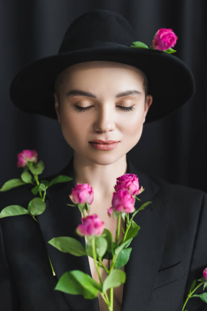 stijlvolle vrouw met gesloten ogen in de buurt wazig roze rozen op zwarte achtergrond - Foto, afbeelding