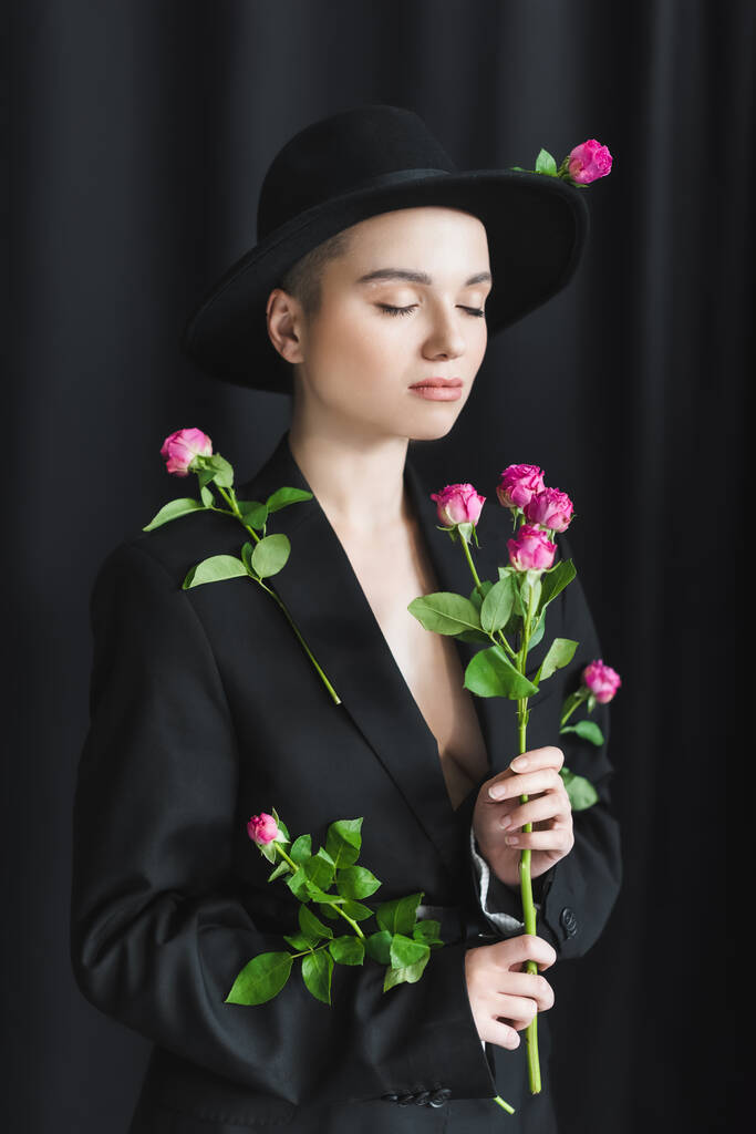 молода жінка в чорному цегляному капелюсі та блістері позує із закритими очима та рожевими трояндами на темному фоні
 - Фото, зображення
