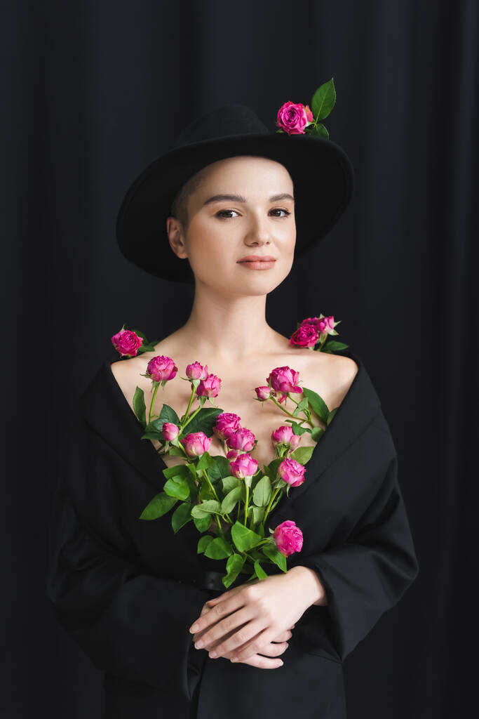 okouzlující žena v černé sako a klobouk pózování s čerstvými růžové růže na tmavém pozadí - Fotografie, Obrázek