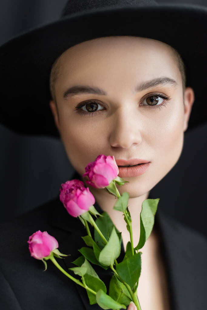 czarująca kobieta w czarnym kapeluszu Fedora patrząc na aparat w pobliżu różowych róż na ciemnym tle - Zdjęcie, obraz