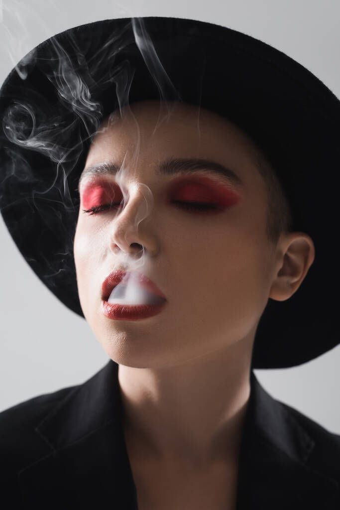 目を閉じて灰色に隔離された煙を吐き出す赤い化粧をした女性 - 写真・画像
