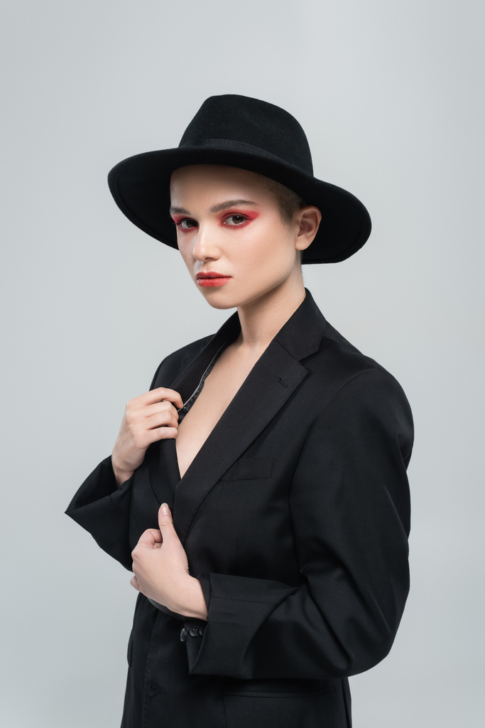 aistillinen nuori nainen lieri hattu koskettaa musta bleiseri katsoessaan kamera eristetty harmaa - Valokuva, kuva