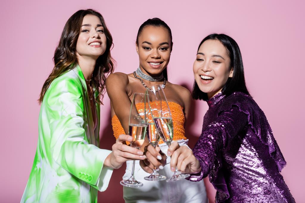 vrolijke interraciale vrouwen in trendy outfits klinkende glazen champagne op roze  - Foto, afbeelding