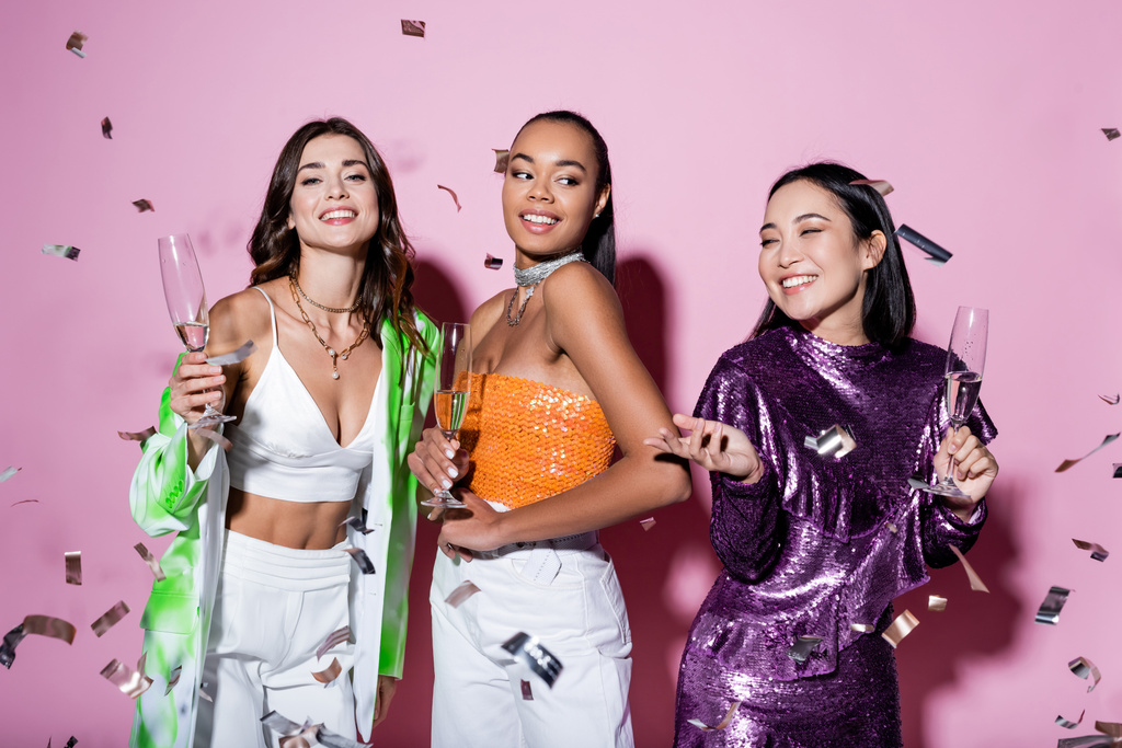 cheerful multiethnic women in trendy outfits holding glasses of champagne near falling confetti on pink  - Photo, Image