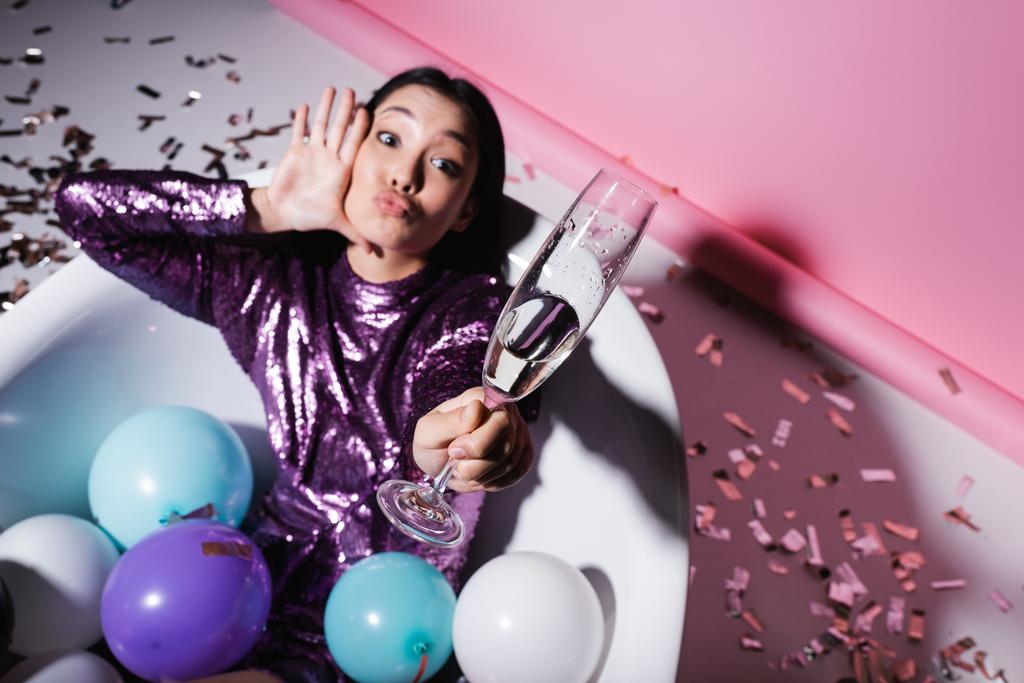 vista de ángulo alto de la mujer asiática haciendo pucheros labios mientras está acostado en la bañera con globos y sosteniendo vidrio borroso de champán - Foto, imagen