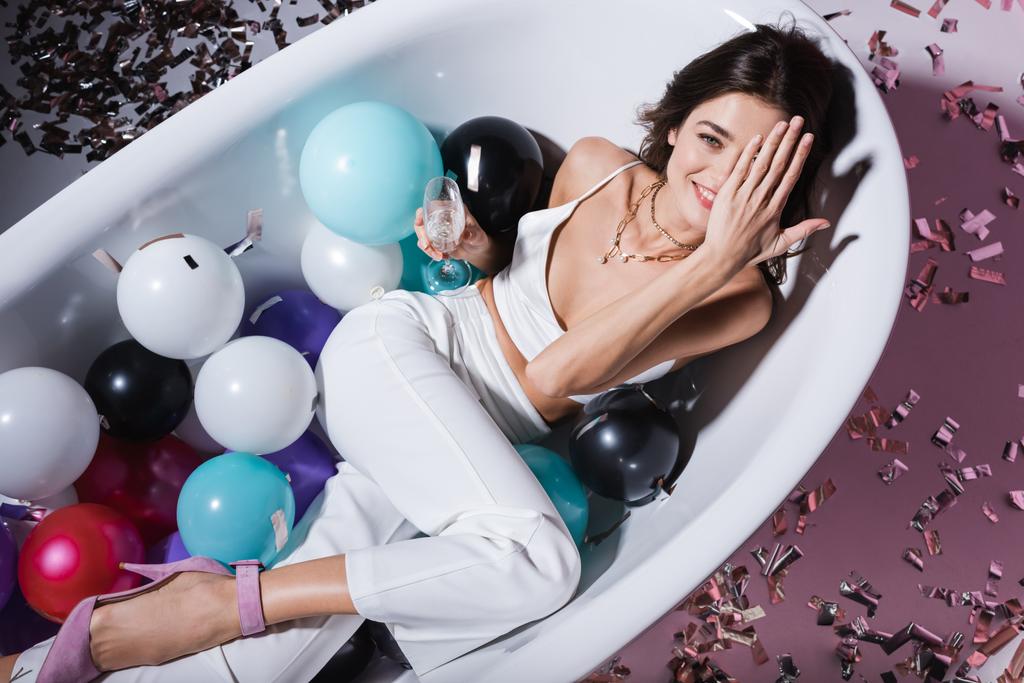 vista superior de la mujer feliz cubriendo el ojo y acostado en la bañera con globos mientras sostiene la copa de champán  - Foto, imagen