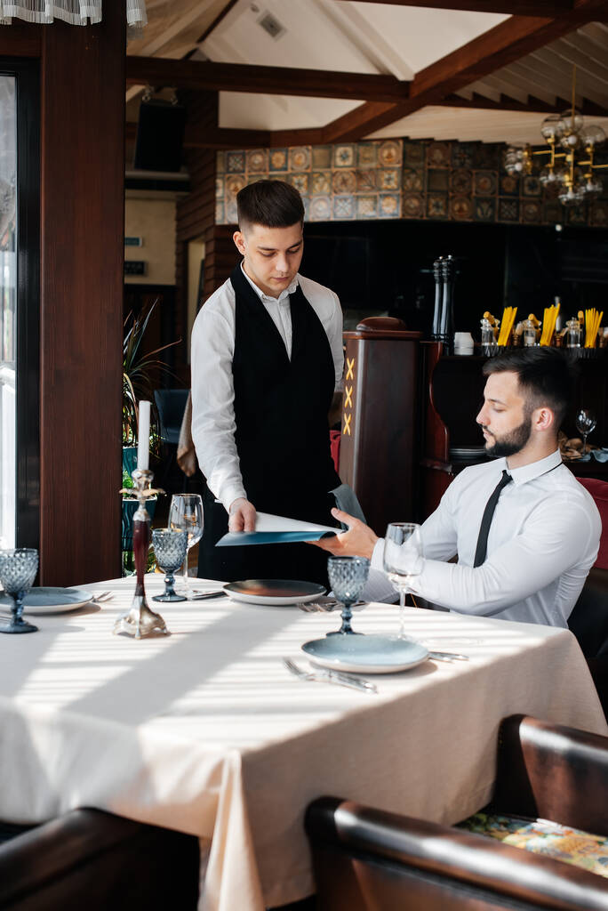 Młody biznesmen w eleganckiej restauracji bada menu i składa zamówienie młodemu kelnerowi w stylowym fartuchu. Obsługa klienta. Obsługa stołu w restauracji. - Zdjęcie, obraz