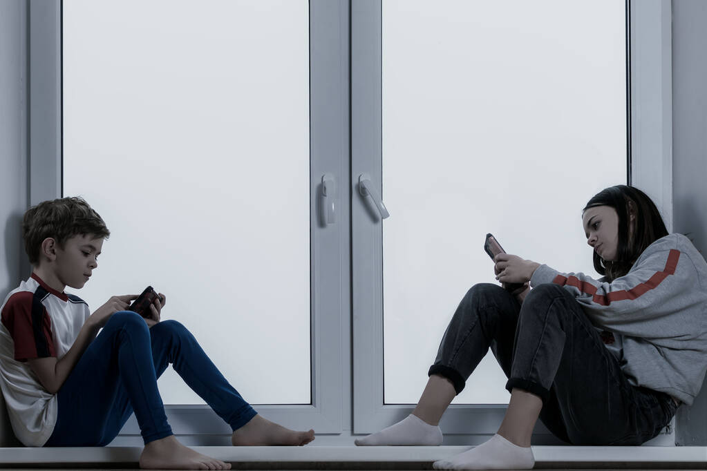 Ein kleiner Junge und seine Schwester sitzen am Fenster und spielen mit ihren Telefonen - Foto, Bild