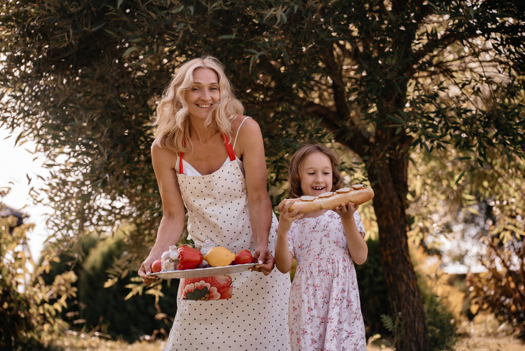 Kleines Mädchen und ihre Mutter in weißen Kleidern mit Obst und Gemüse in den Händen auf dem Hintergrund von Bäumen. Sommerpicknick in der Natur. Mutter und Tochter amüsieren sich gemeinsam im Park. - Foto, Bild