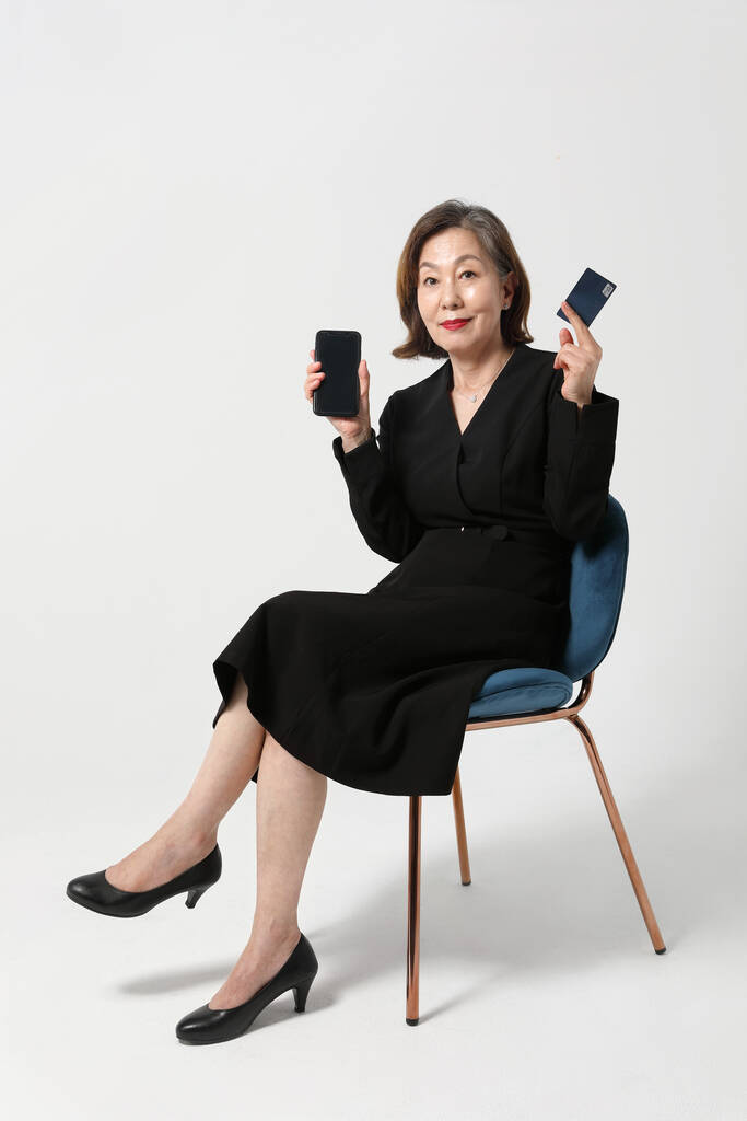 ファッショナブルなアジア系韓国人のシニア女性で、クレジットカードとスマートフォンを持って椅子に座って - 写真・画像