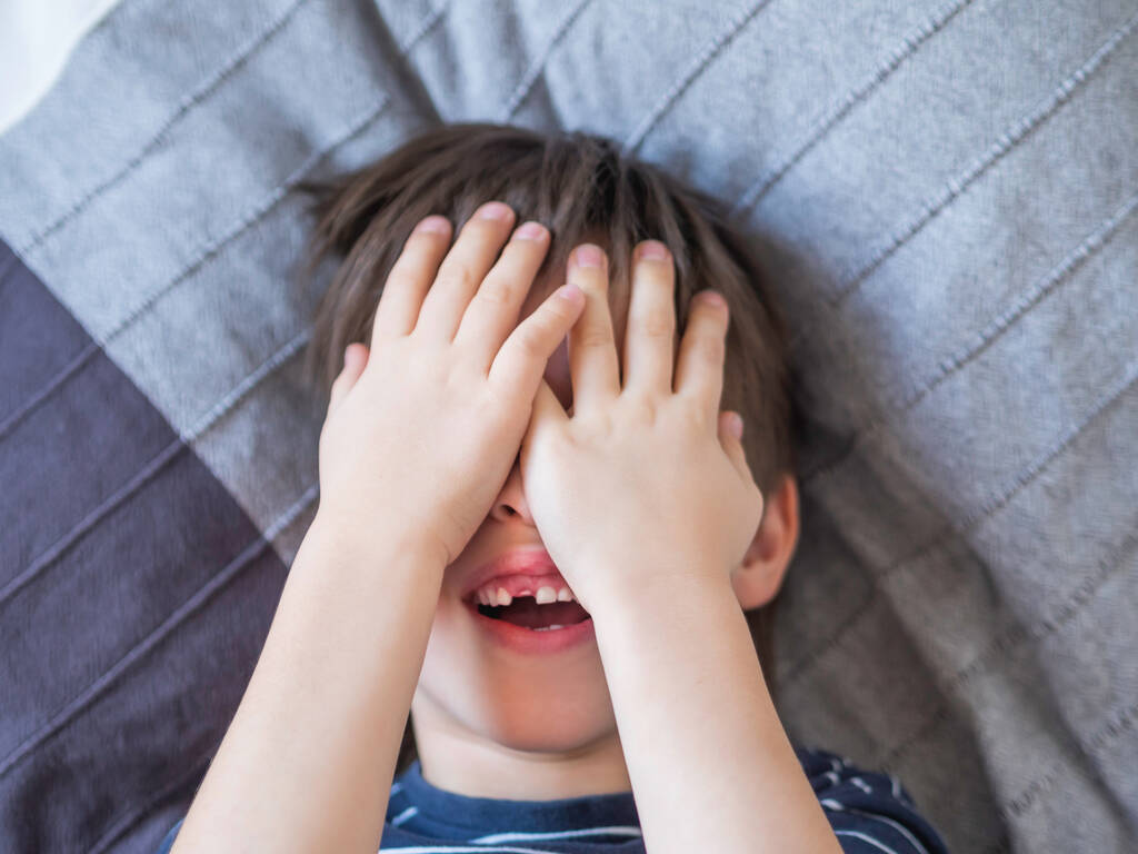Lachend kind sluit ogen met handen maar toont gat in rij tanden in zijn mond. Een snijtand viel er net uit. Close up foto van tandvlees voor tandarts.  - Foto, afbeelding
