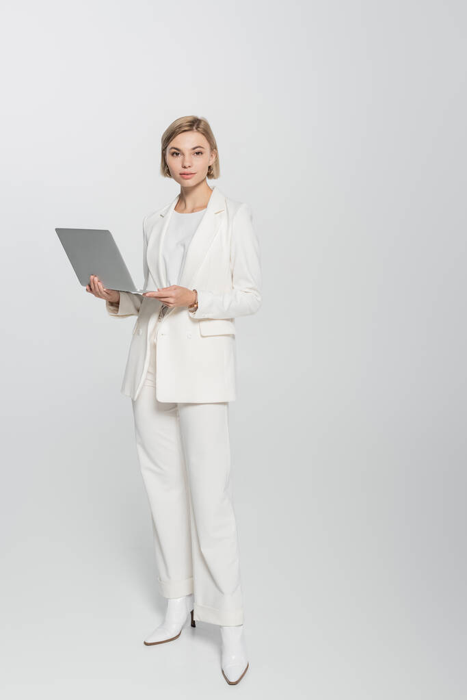 Повна довжина молодої бізнес-леді в костюмі тримає ноутбук і дивиться на камеру на сірому фоні
  - Фото, зображення