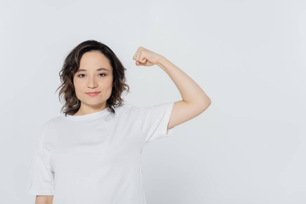 Молодая женщина в белой футболке показывает мышцы, изолированные на сером  - Фото, изображение
