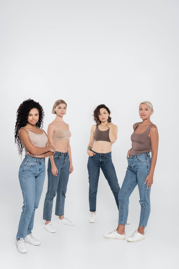 Multiethnische Frauen in Jeans posieren und blicken in die Kamera auf grauem Hintergrund  - Foto, Bild