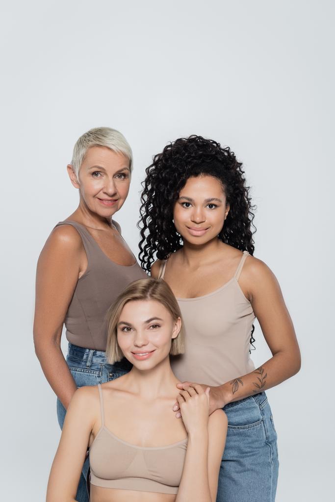 Smiling interracial women looking at camera near young blonde friend isolated on grey  - Photo, Image