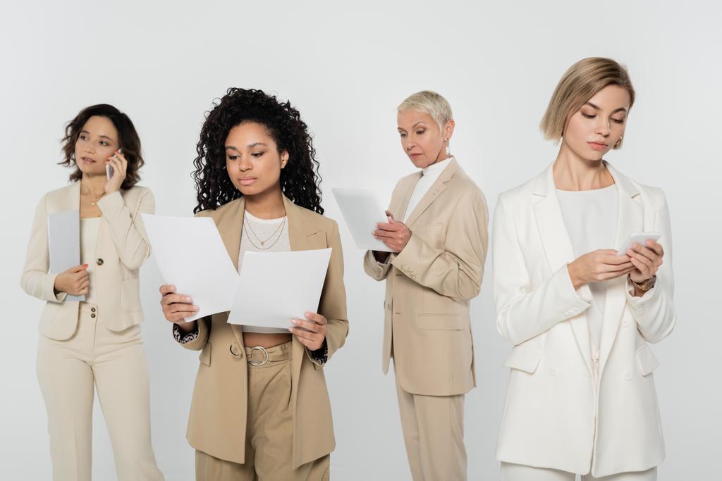 Multiethnic businesswomen holding papers and using devices isolated on grey  - Photo, Image
