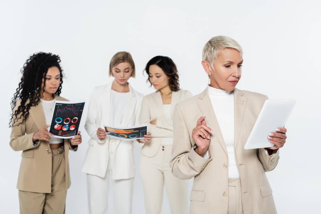Літня бізнес-леді використовує цифровий планшет поблизу розмитих міжрасових колег з паперами ізольовані на сірому
  - Фото, зображення
