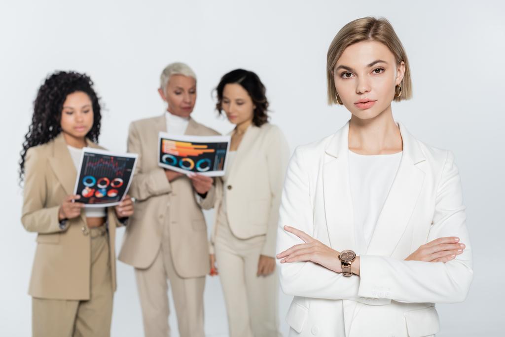 Femme d'affaires blonde regardant la caméra près de collègues interracial flous avec des papiers isolés sur gris  - Photo, image
