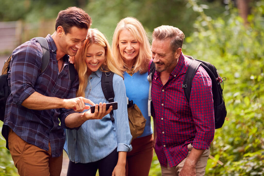 Группа друзей, путешествующих в сельской местности, рассматривая фотографии по телефону, когда они идут по тропе - Фото, изображение