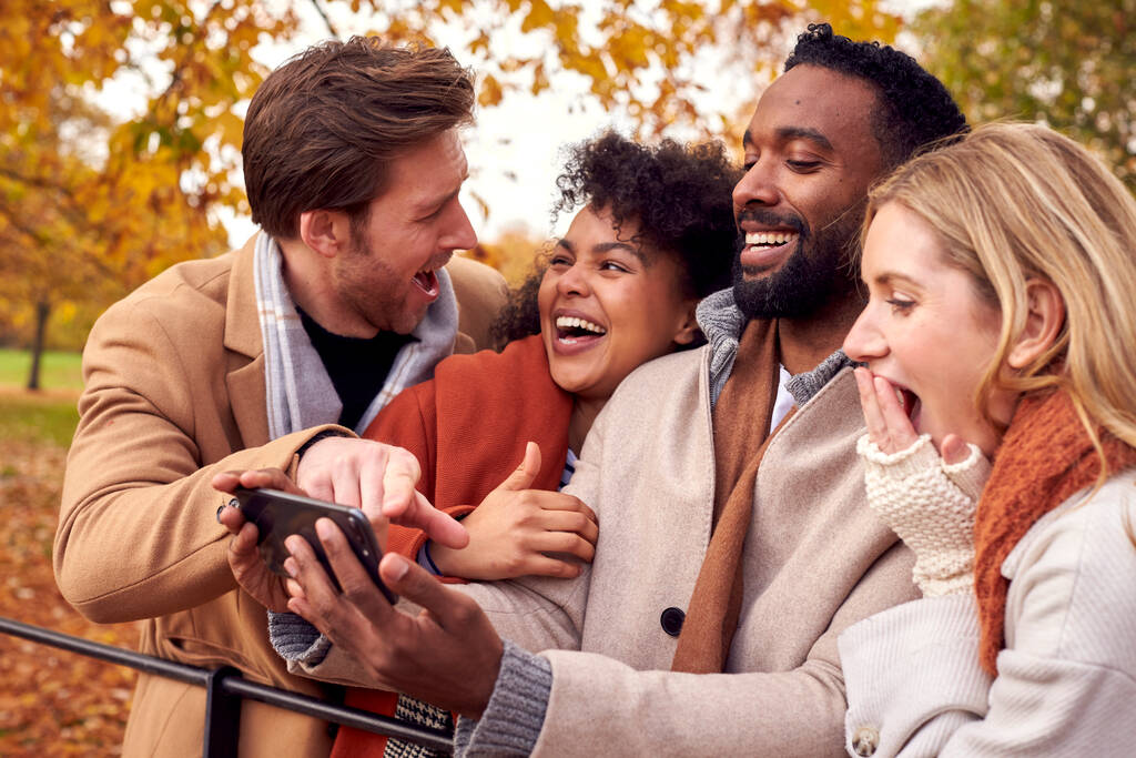Grupo de amigos ao ar livre vestindo casacos e cachecóis olhando para fotos no telefone no parque de outono - Foto, Imagem