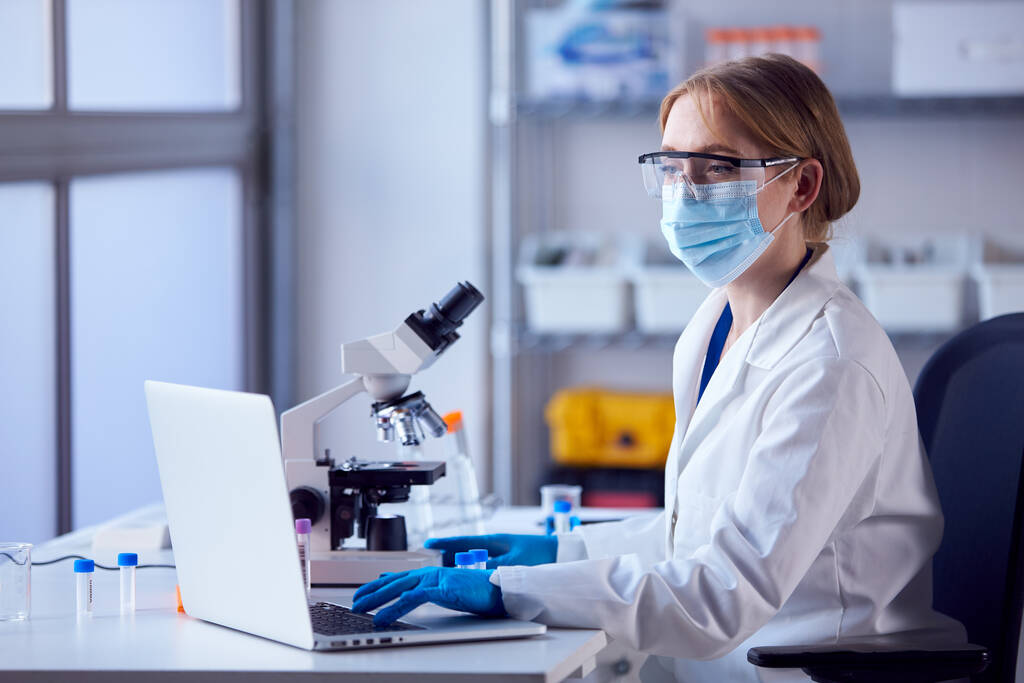 Kobiecy pracownik laboratorium noszący fartuch laboratoryjny Analizujący próbki w laboratorium z laptopem i mikroskopem - Zdjęcie, obraz