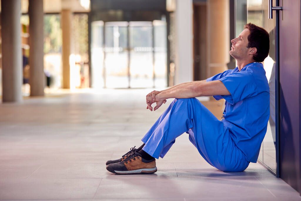 Втомлений і перероблений медичний працівник сидить на підлозі лікарняного коридору під час перерви
 - Фото, зображення