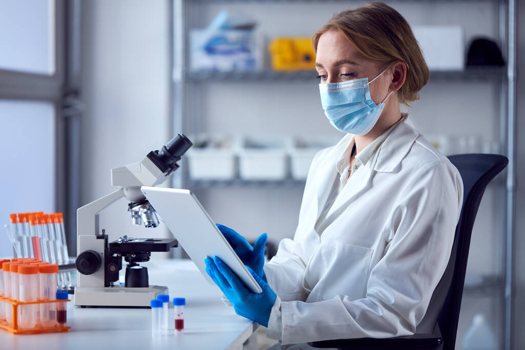 Travailleur de laboratoire féminin portant un manteau de laboratoire travaillant sur le vaccin Covid-19 avec tablette numérique et microscope - Photo, image