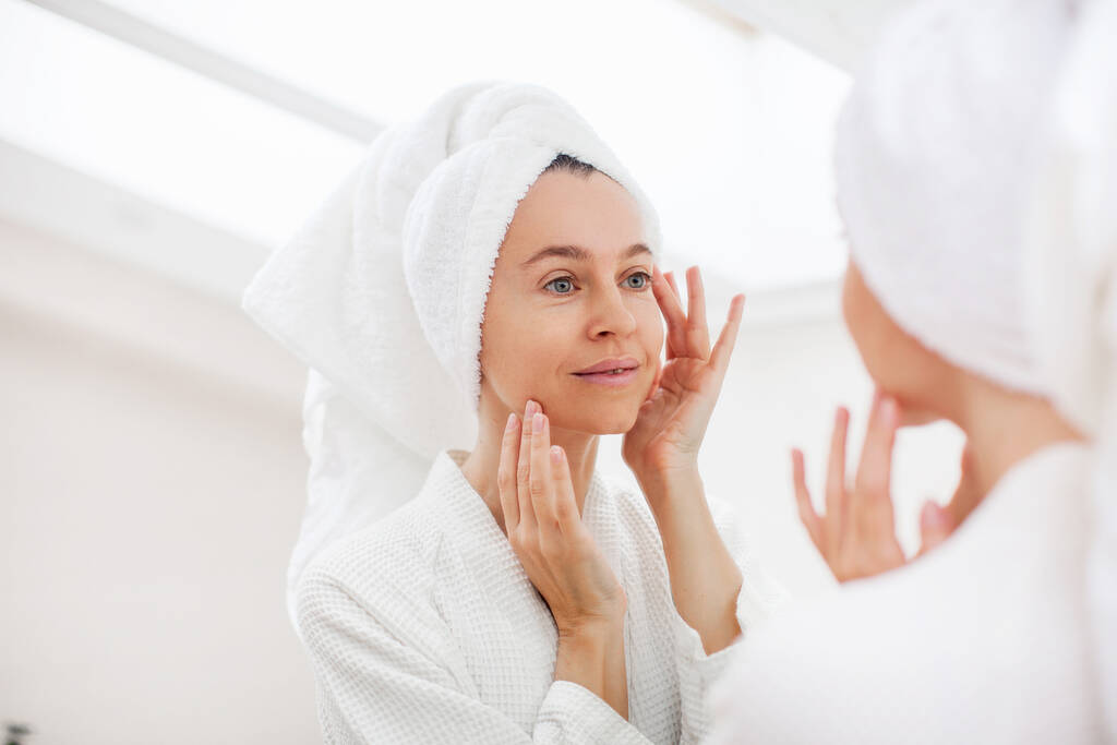 Κοντινό πορτρέτο της πανέμορφης ευτυχισμένης μεσήλικας γυναίκας που κοιτάζει τον καθρέφτη αγγίζοντας το δέρμα της απολαμβάνοντας τη θεραπεία για το ξηρό δέρμα. Διαφήμιση προϊόντων περιποίησης δέρματος κατά της γήρανσης - Φωτογραφία, εικόνα