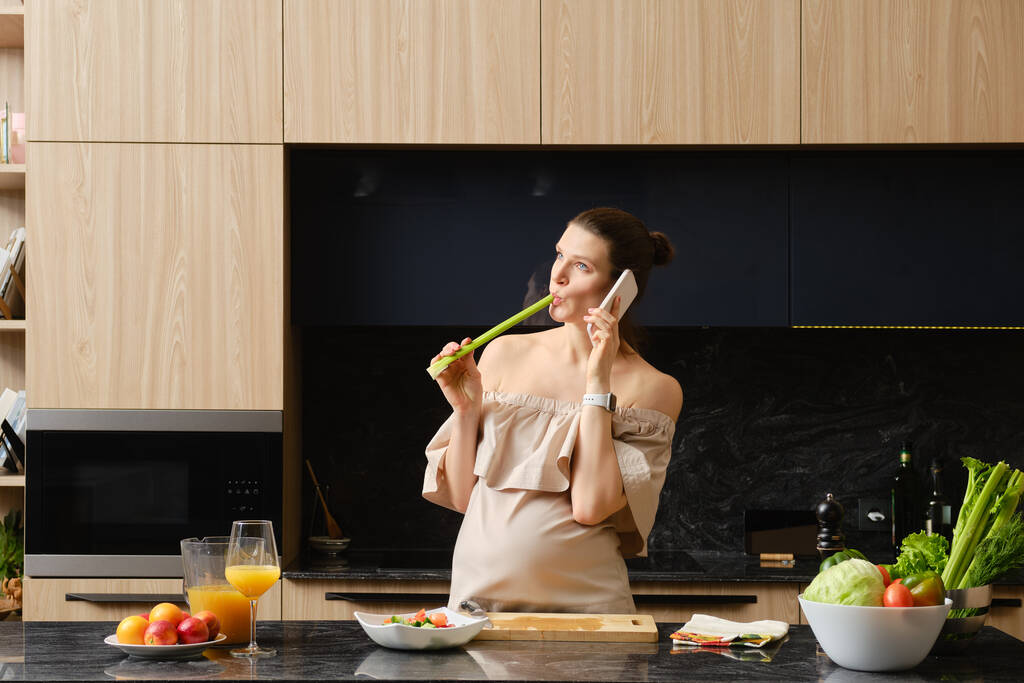Těhotná žena okusuje celer stonek a mluví po telefonu při přípravě jídla v kuchyni - Fotografie, Obrázek