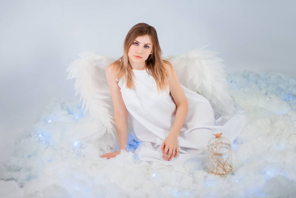 Una chica atractiva en una túnica blanca con grandes alas blancas detrás de su espalda posa mientras está sentada en nubes blancas y luminosas. - Foto, imagen