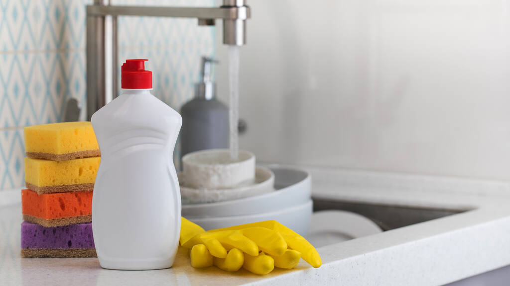 Ένα λευκό μπουκάλι με τζελ πιάτων, σφουγγάρια και λαστιχένια γάντια στο φόντο ενός νεροχύτη με βρώμικα πιάτα. Αντιγραφή χώρου - Φωτογραφία, εικόνα