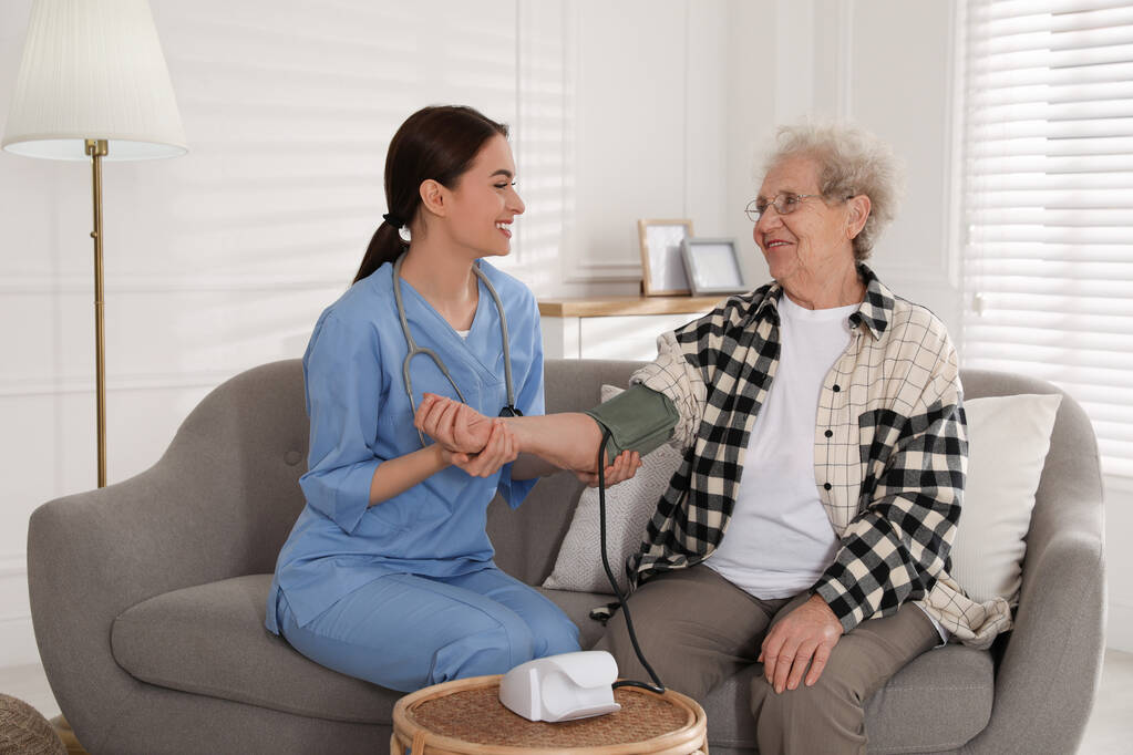 Cuidador joven midiendo la presión arterial de la mujer mayor en la habitación. Servicio de atención médica domiciliaria - Foto, imagen
