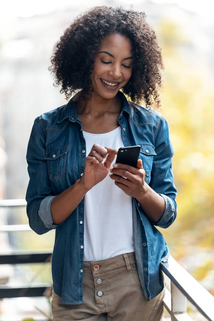 Foto de mujer alegre usando su teléfono inteligente mientras envía mensajes de pie en el balcón en casa. - Foto, Imagen