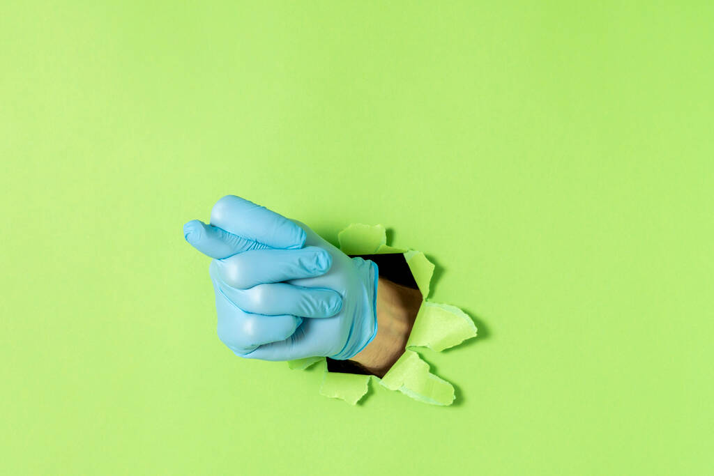 Fig as a hand gesture, denoting humiliation or denial. Male hand in blue latex glove sticks out of a torn hole in green paper. Copy space. - Photo, Image
