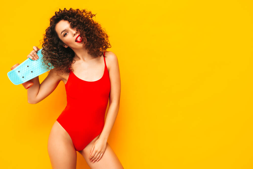 Jovem bela mulher sorridente posando perto da parede amarela no studio.Sexy modelo em roupa de banho de banho vermelho.Mulher positiva com cachos penteado. Segurando penny skateboard.Happy e alegre - Foto, Imagem