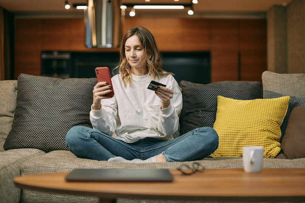 Jonge vrouw met bankkaart, invoeren van betalingsinformatie in de telefoon, online zittend op de bank thuis kopen  - Foto, afbeelding