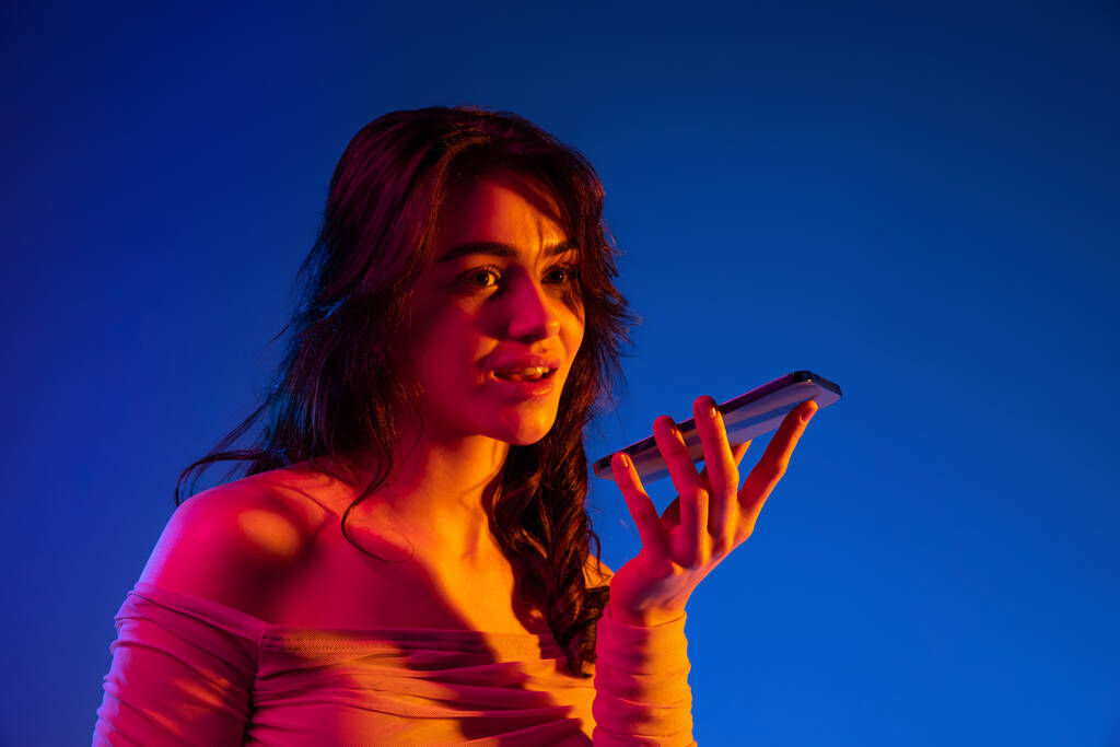 Notícias. Retrato de menina emocional jovem, estudante com cabelos longos escuros usando gadget isolado em fundo azul escuro em luz de néon. Conceito de emoções - Foto, Imagem