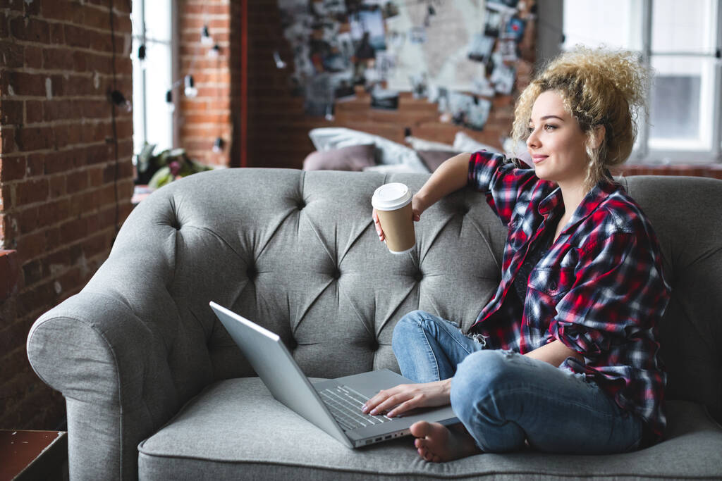Mosolygó gyönyörű fehér nő göndör haj kanapén laptop és kávé. Szabadúszó, távoli munkavégzés otthonról. - Fotó, kép