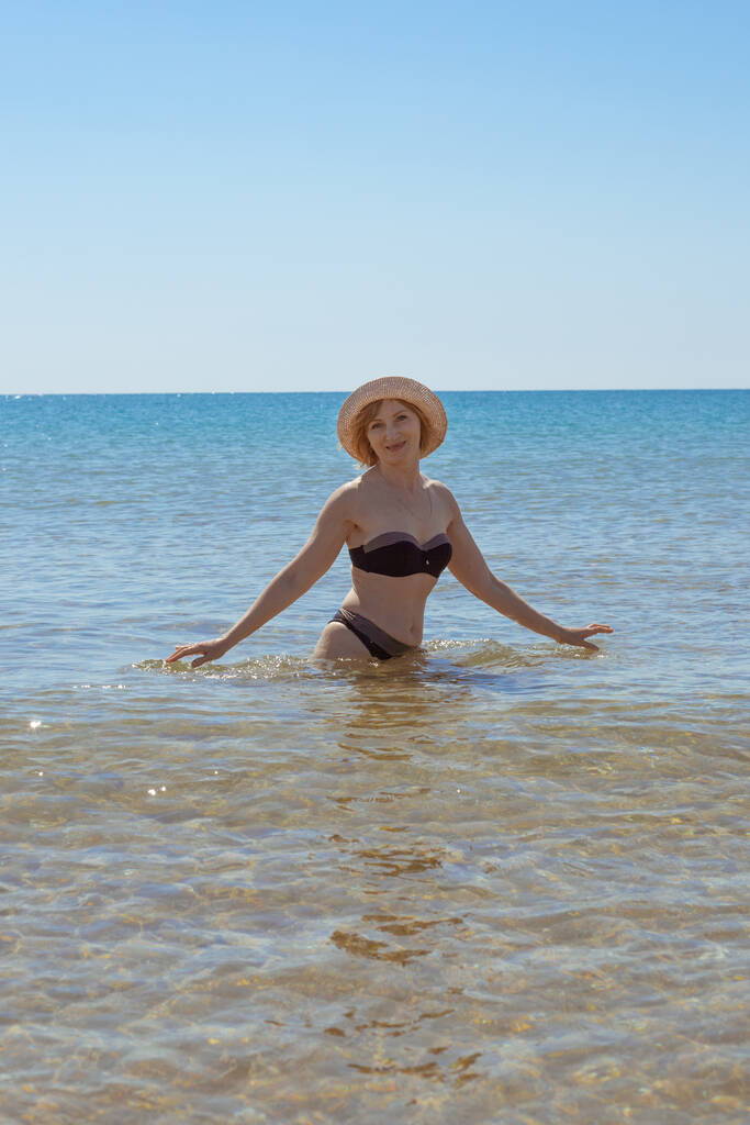 Mujer europea en traje de baño y sombrero de cintura en el mar sonríe y mira a la cámara. Vacaciones en la playa. - Foto, imagen