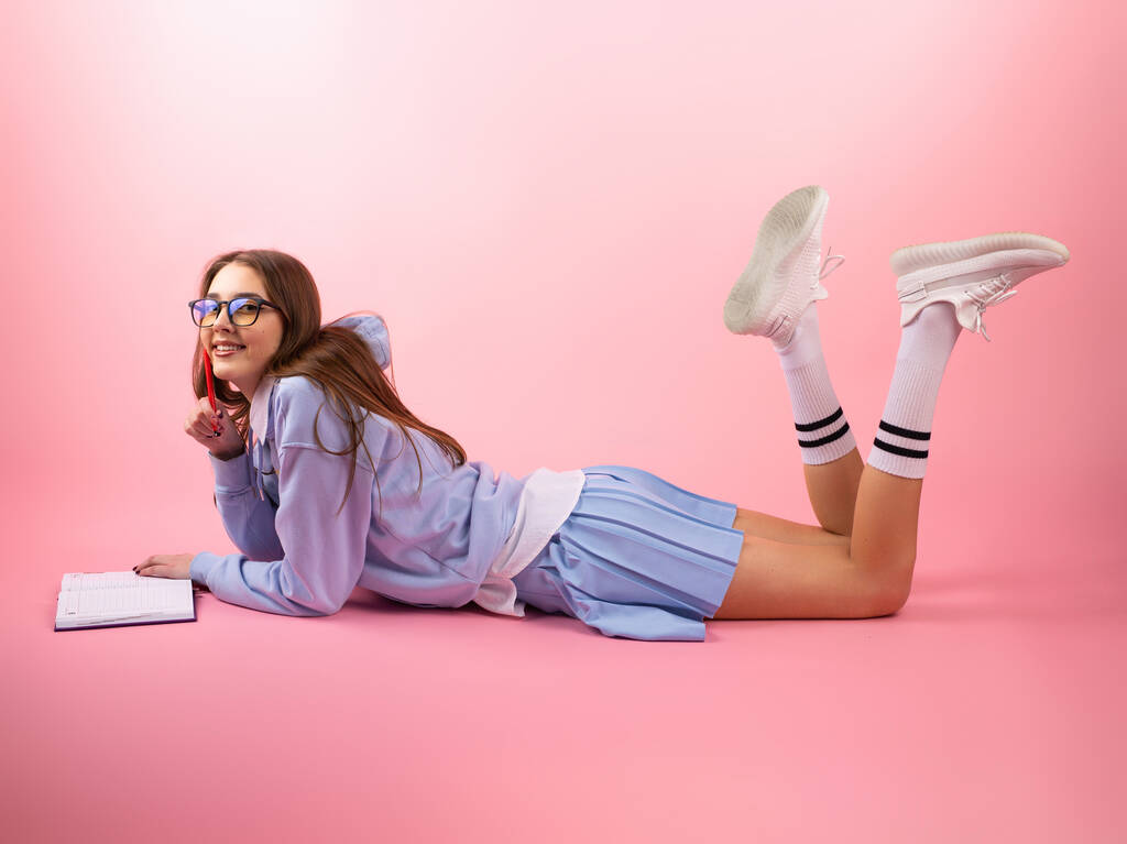 Satisfeito menina estudante bonita vestindo uniforme azul e óculos deitados no chão em seu estômago e levantou as pernas para cima dobrando os joelhos e lendo o livro. - Foto, Imagem