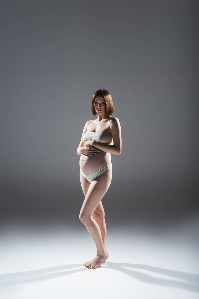 İç çamaşırlı çıplak ayaklı hamile kadın gri arka planda kameraya bakıyor. - Fotoğraf, Görsel
