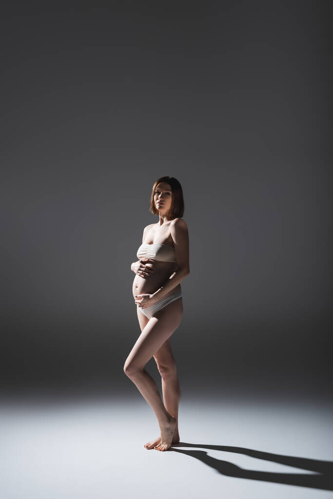 Ξυπόλητη έγκυος γυναίκα με εσώρουχα στέκεται σε γκρι φόντο με σκιά  - Φωτογραφία, εικόνα