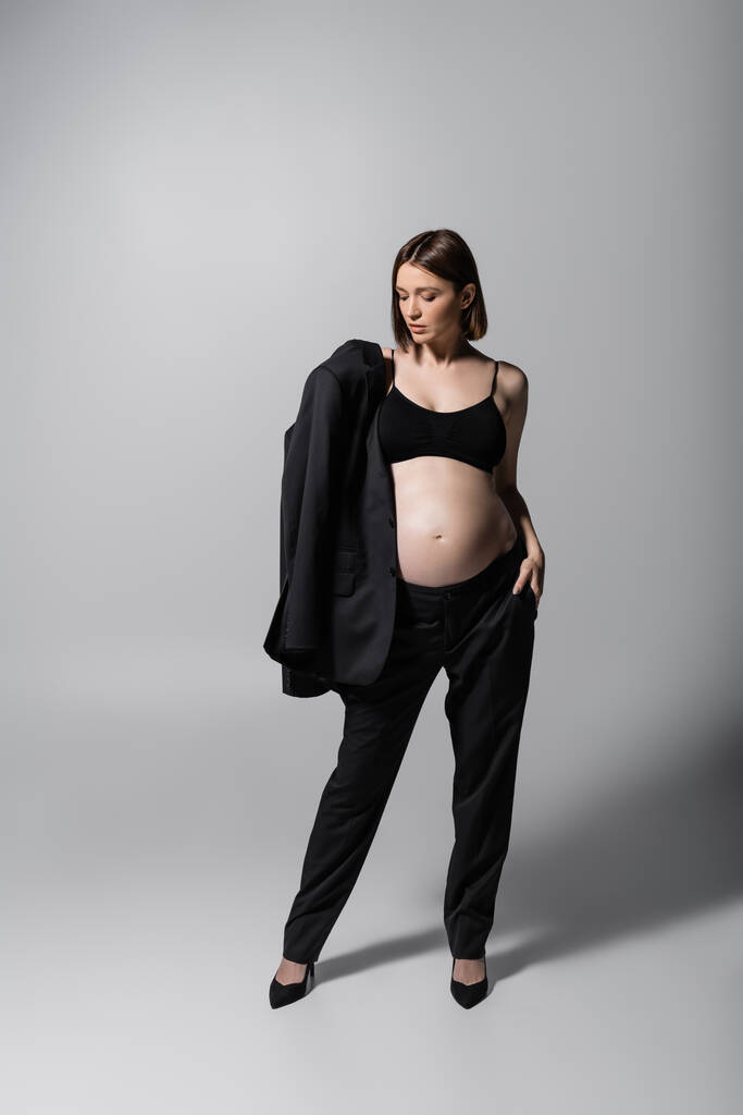 Elegante modelo embarazada en la parte superior y tacones que sostiene la chaqueta sobre fondo gris - Foto, imagen
