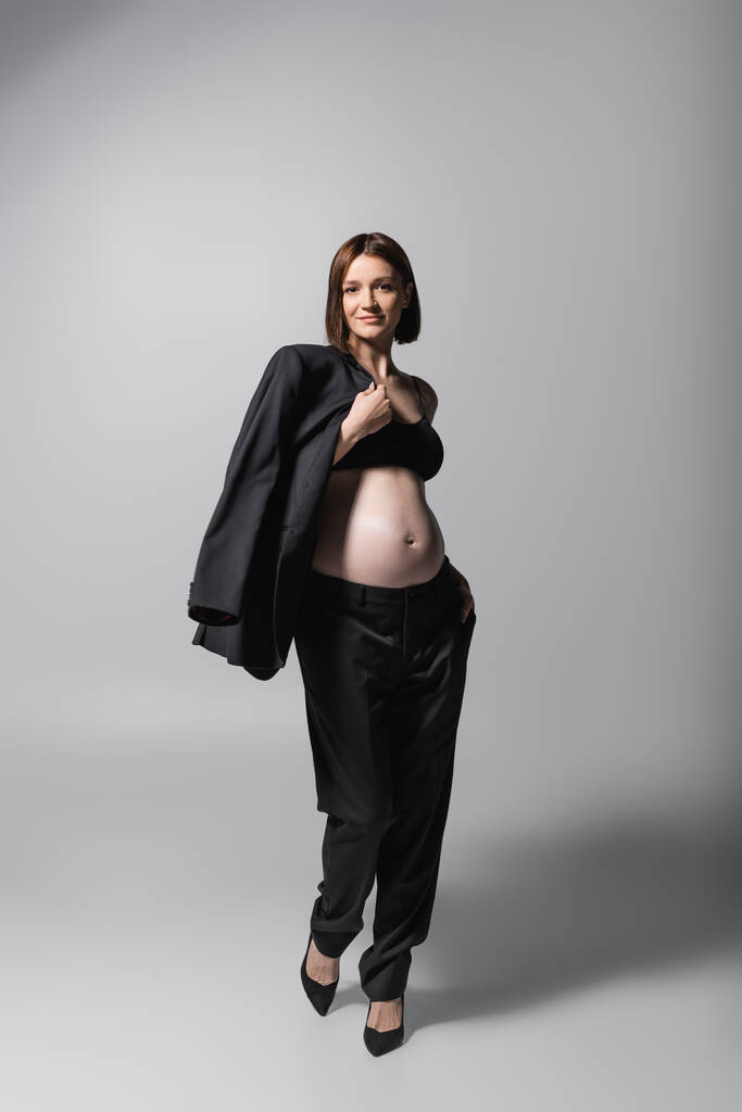 Χαμογελώντας έγκυος γυναίκα στην κορυφή κρατώντας σακάκι σε γκρι φόντο - Φωτογραφία, εικόνα